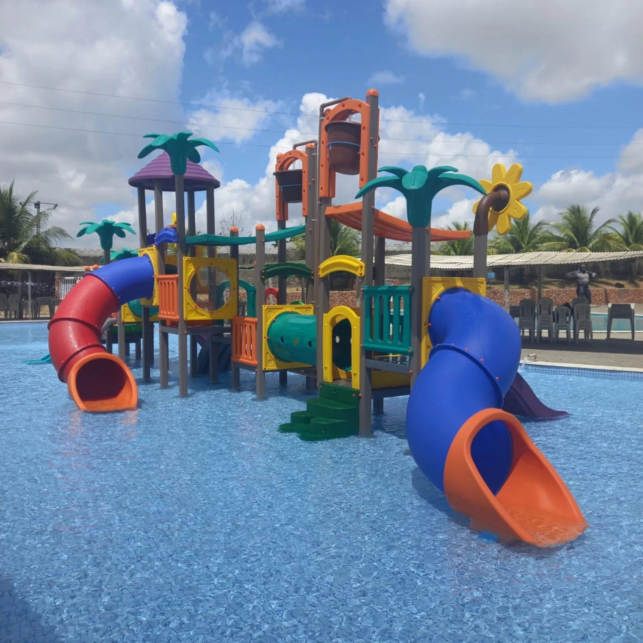 Parque infantil aquático com escorregadores coloridos em piscina, ao ar livre, com palmeiras e céu azul ao fundo, em área de lazer.